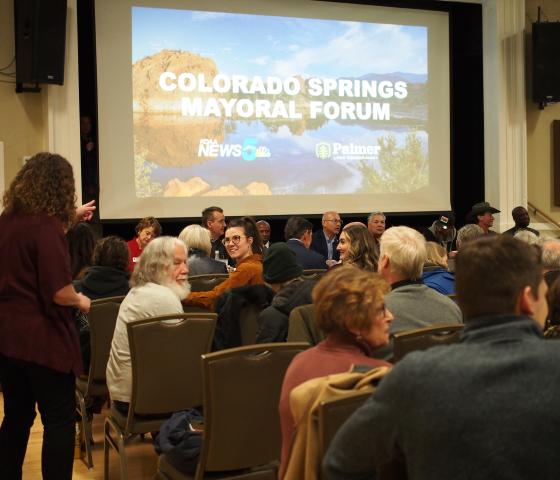 Attendees sitting facing a stage with mayoral candidates. A large screen behind the candidates reads "Colorado Springs Mayoral Forum"