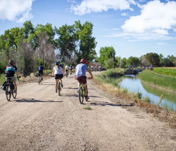 Cyclists bike along an irrigation ditch and farm lands.