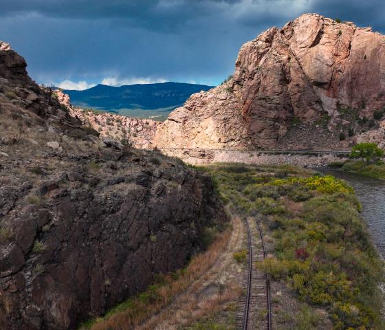 Light illuminates a rocky crag in a deep canyon with train tracks and a river at the bottom. 