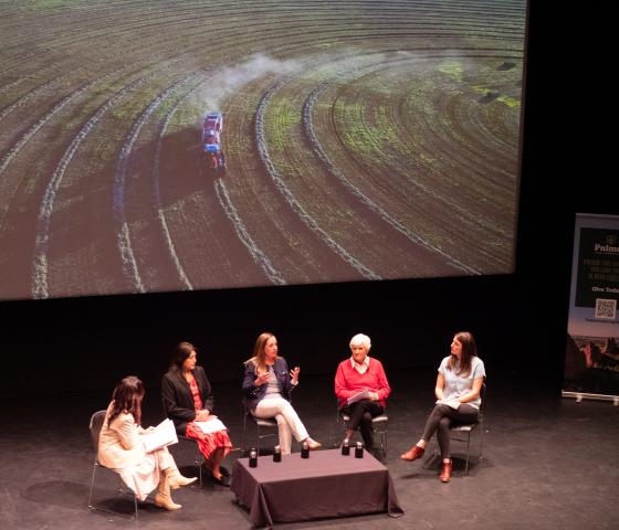 Five women sit in a semi-circle on a dark stage with a large projector image of a farm in the background. 