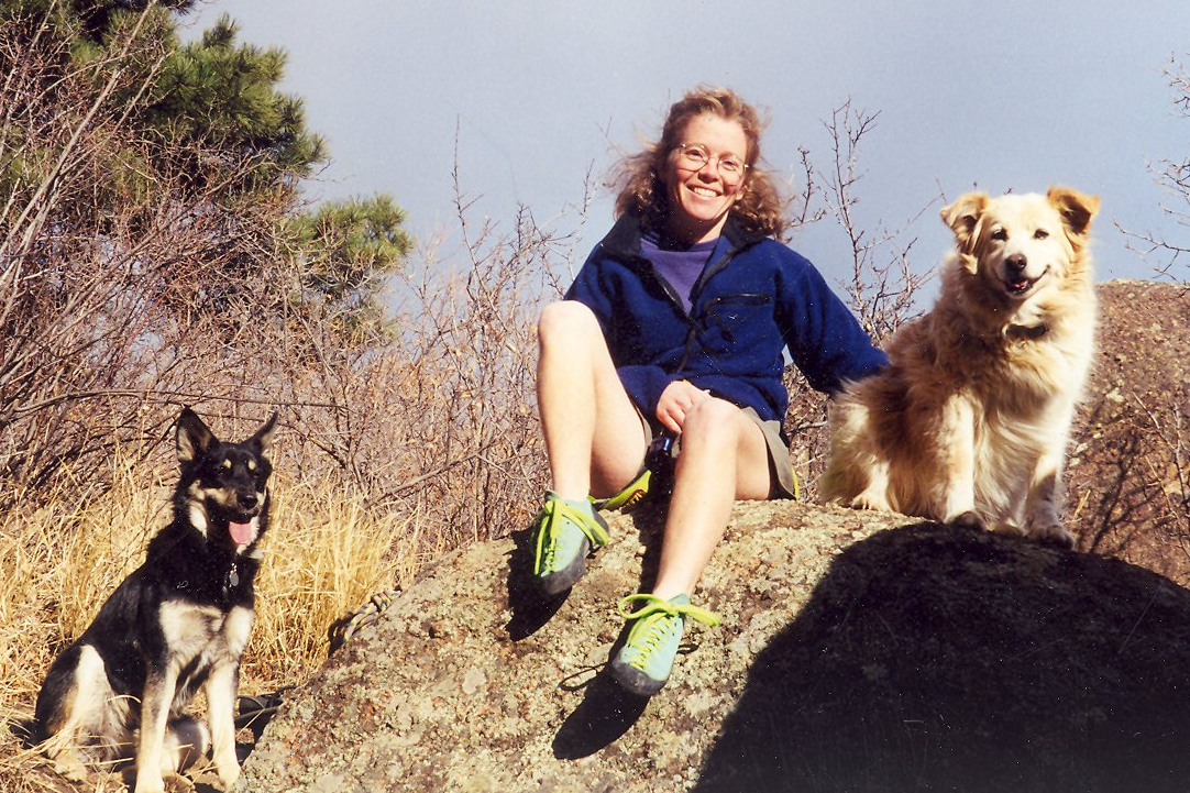 A young women with blond hair and circular glasses wearing a blue fleece and khakis shorts sits on a sun lit rock with two dogs. 