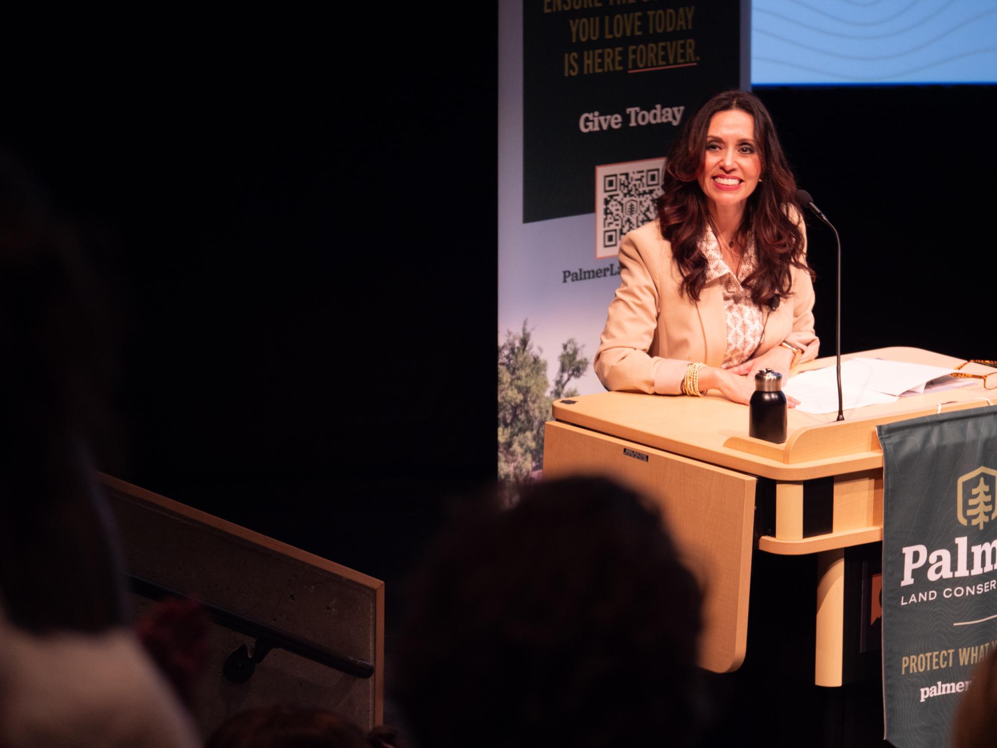 Women with long brown hair smiles at a well-lit podium on a dark stage.